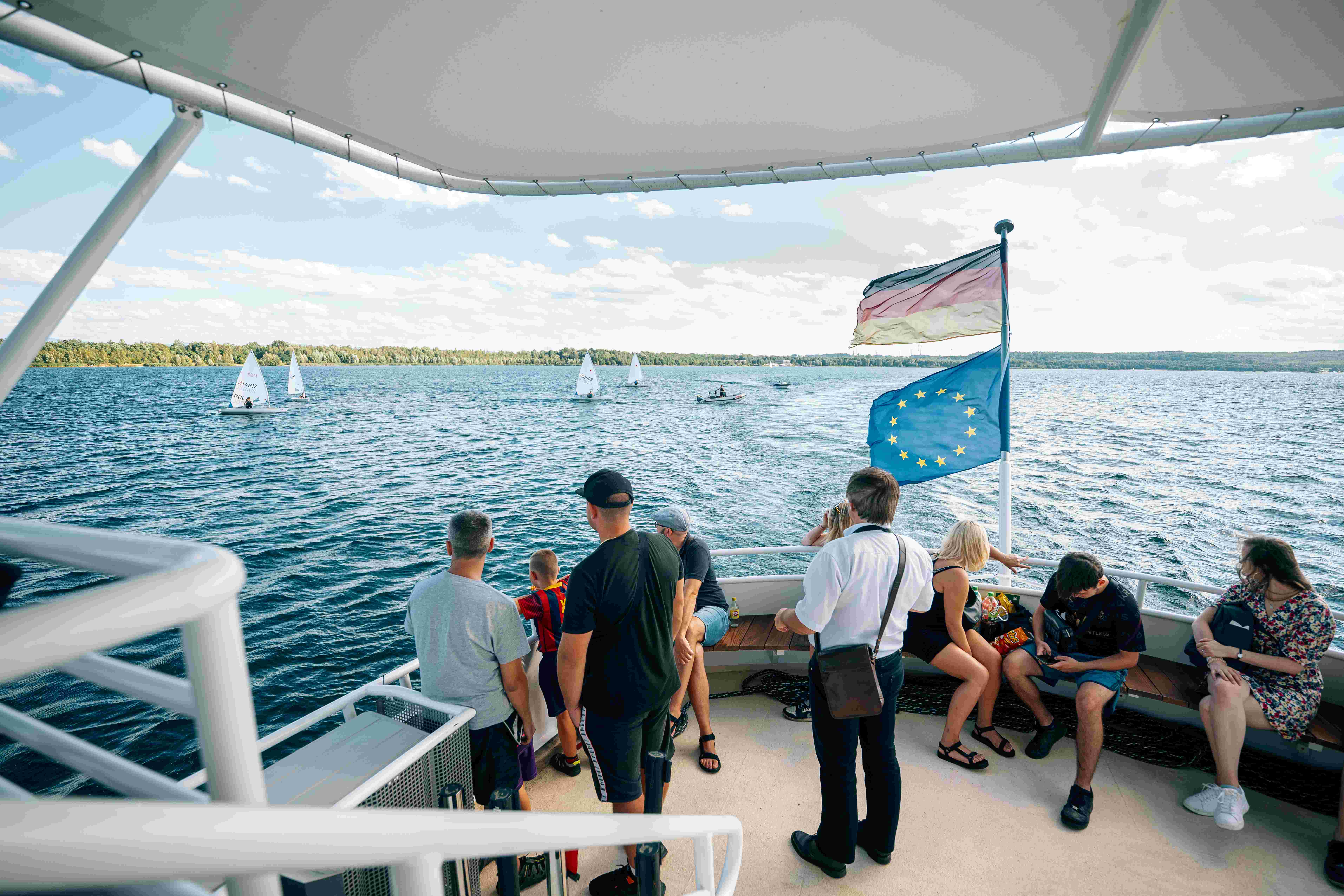 Pasażerowie stoją na dziobie statku i patrzą na jezioro.